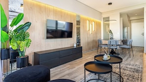 Uma sala de estar moderna com televisão de tela plana, painéis de madeira, um rack de entretenimento preto e uma área de jantar com mesa e cadeiras.