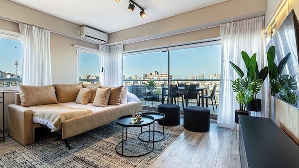 Sala de estar moderna com sofá, mesa de centro e cadeiras, grandes janelas com vista para a cidade e uma varanda com plantas.