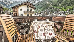 Una terraza de madera con una mesa y sillas con vistas a un pueblo de montaña con un chalet y una iglesia.