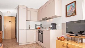 Una cocina compacta con armarios blancos, un horno integrado, una cafetera y un televisor sobre un soporte de madera.