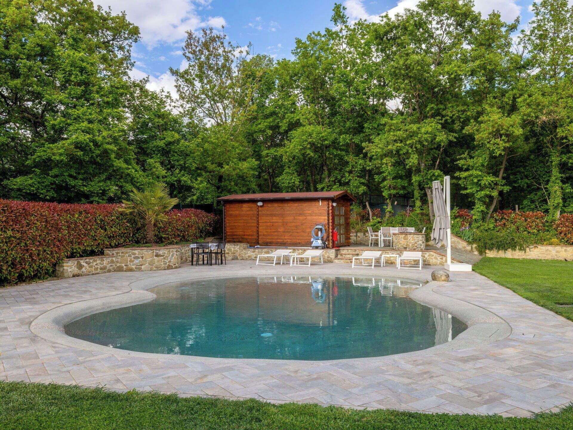 A swimming pool with a wooden deck and lounge chairs, surrounded by a stone patio and lush greenery.