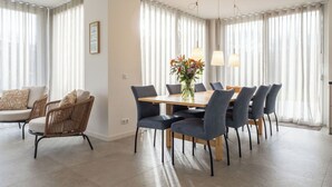 Un área de comedor moderna con una mesa y sillas de madera, ventanas amplias con cortinas ligeras y un jarrón con flores sobre la mesa.