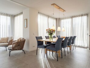 Ein moderner Essbereich mit Holztisch und -stühlen, großen Fenstern mit leichten Vorhängen und einem Blumenstrauß auf dem Tisch.