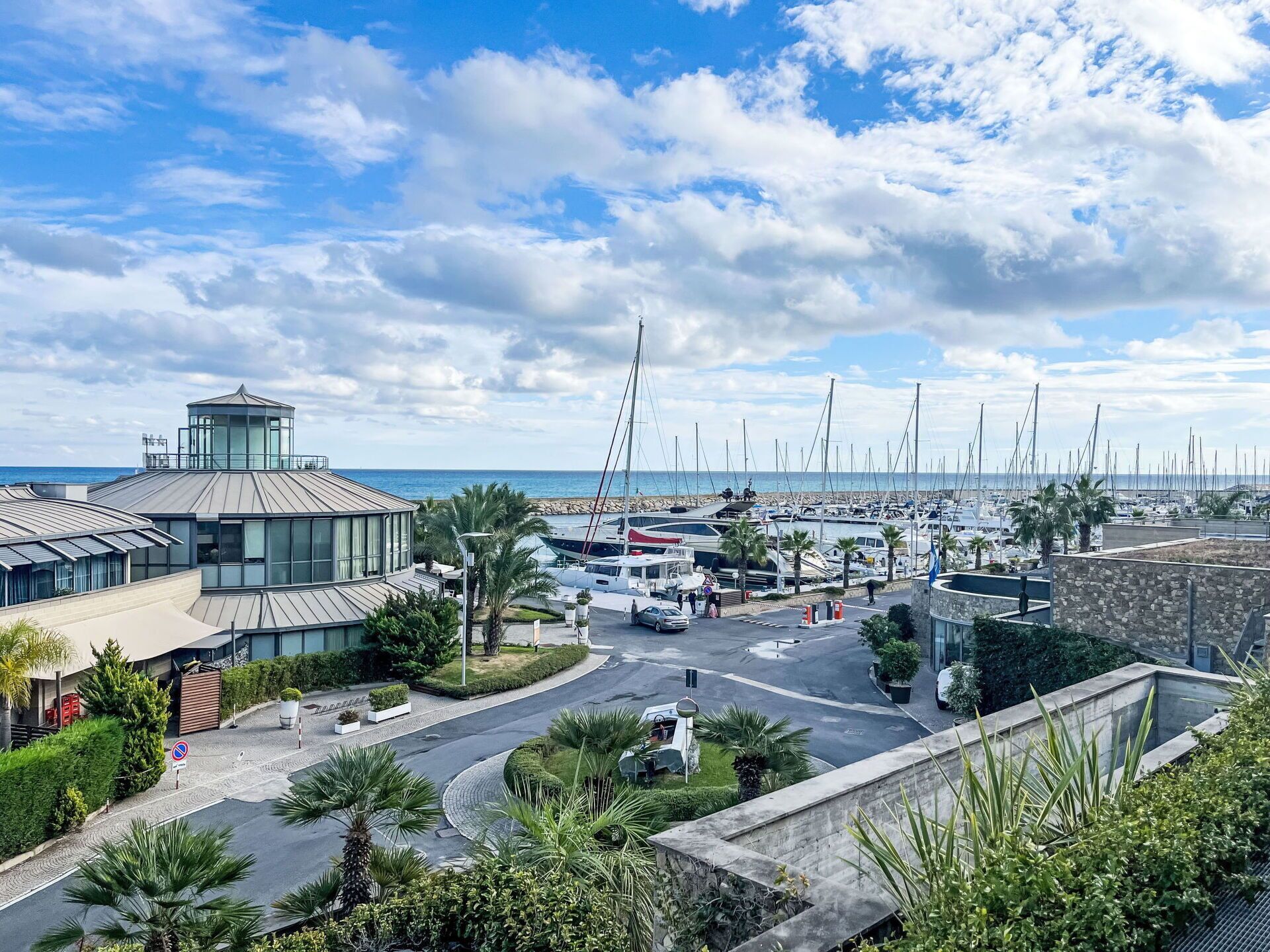 Un port de plaisance avec des bateaux, un bâtiment moderne doté d’une façade en verre et une vue dégagée sur la mer.