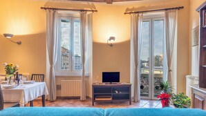 Un salon chaleureux avec un canapé, une table à manger dressée avec des assiettes et un vase de fleurs, un téléviseur posé sur un meuble et de grandes fenêtres avec des rideaux.