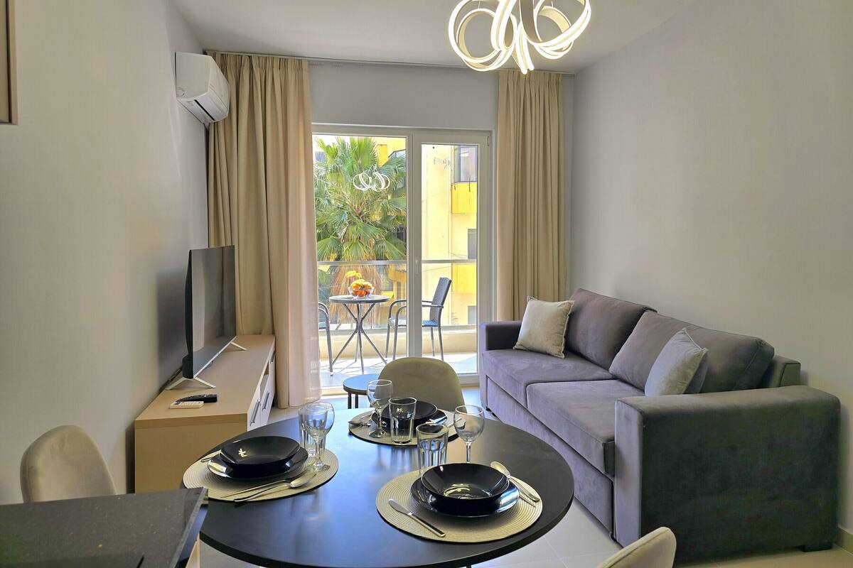 Een moderne woonkamer met een eettafel gedekt voor twee personen, een bank, een televisie en een balkon met uitzicht op een palmboom.