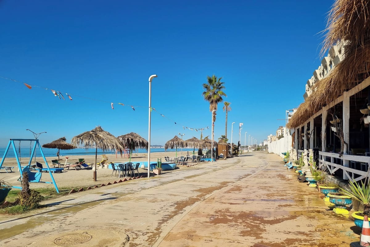 Een promenade aan het strand met rieten parasols, palmbomen en terrasmeubels.