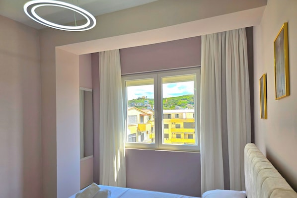 Hotelkamer met een bed, een raam met gordijnen, uitzicht op gebouwen en een ronde lamp.