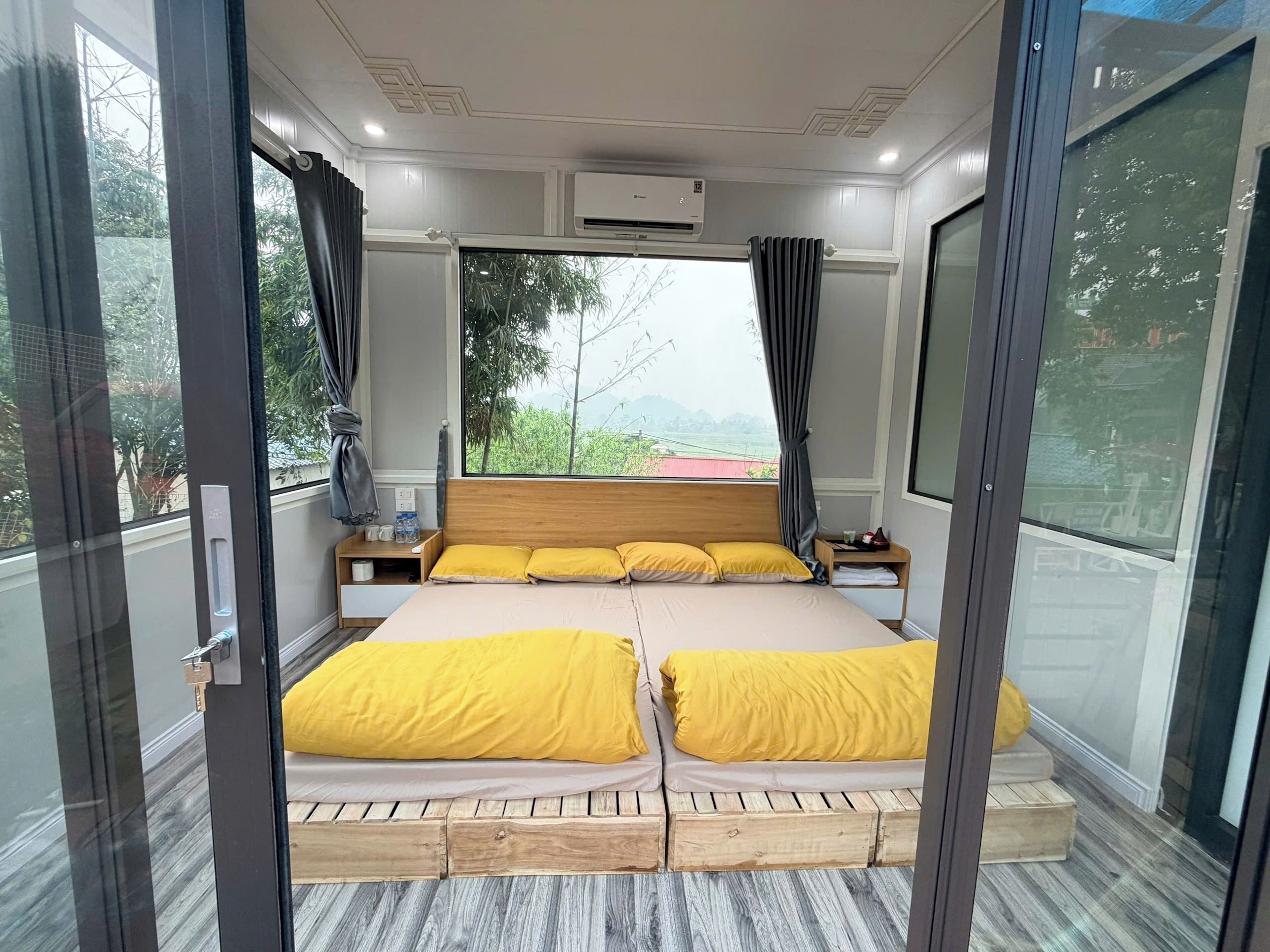 Un dormitorio moderno con una cama de madera, dos almohadas amarillas, una mesita de noche y vistas a zonas verdes.