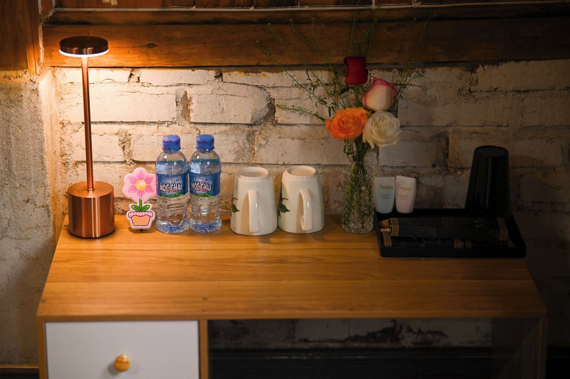 A wooden table with a lamp, water bottles, mugs, a vase with flowers, and a black tray with items.