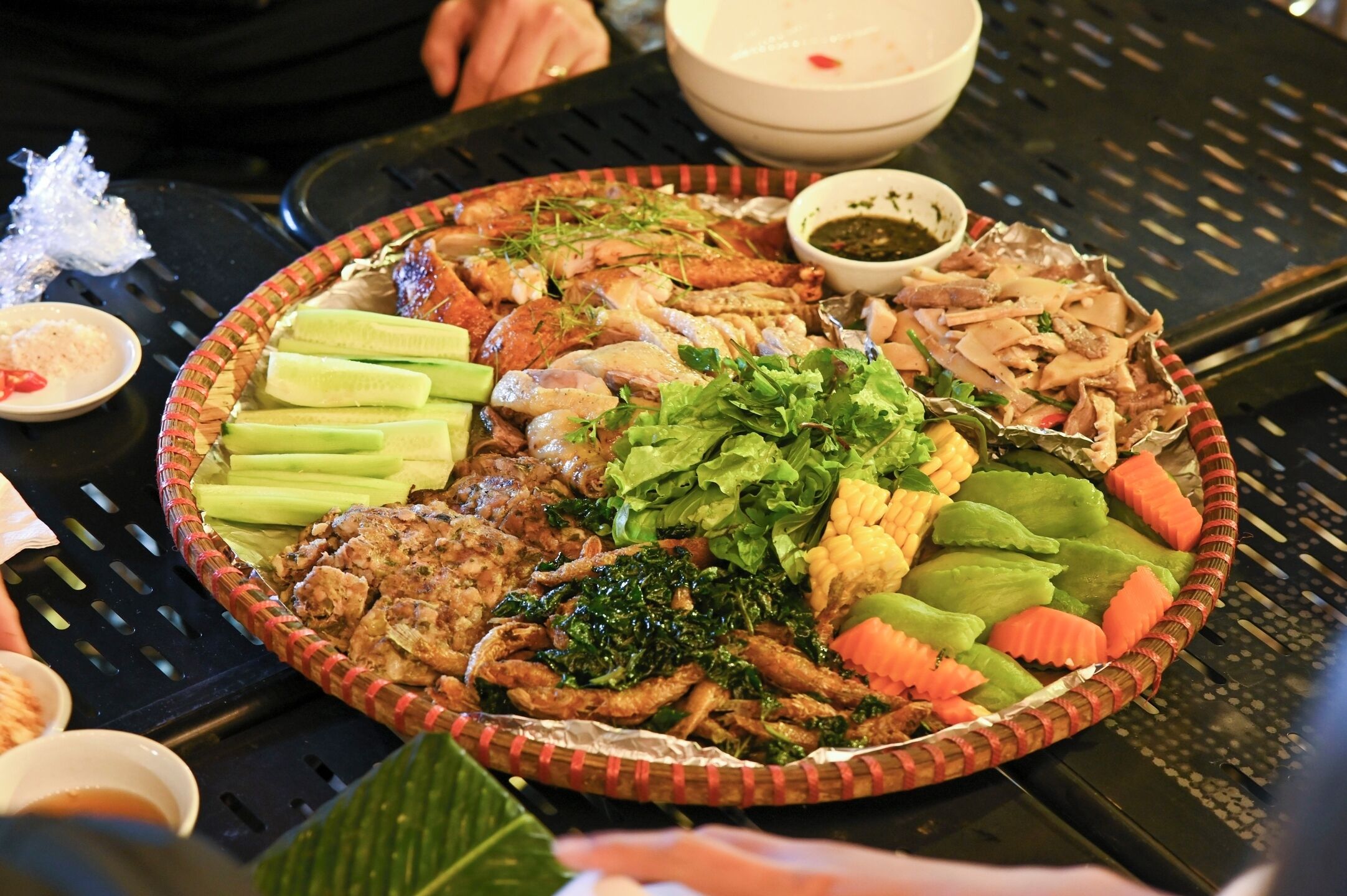 A large, round, woven basket filled with various grilled meats, vegetables, and greens, accompanied by small bowls of sauces.