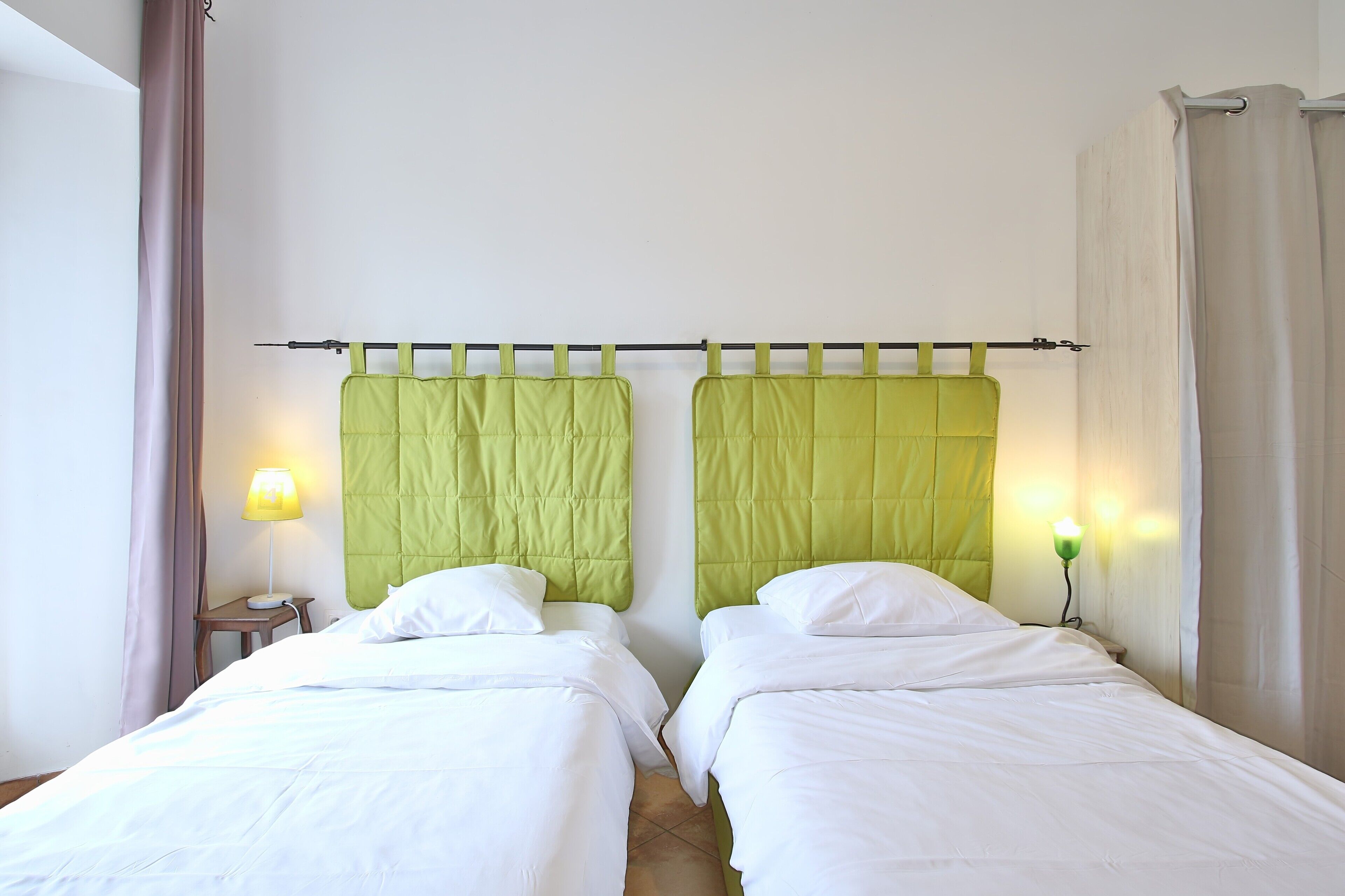 Ein Hotelzimmer mit zwei Einzelbetten, einem Nachttisch mit Lampe und einem Fenster mit Vorhängen.