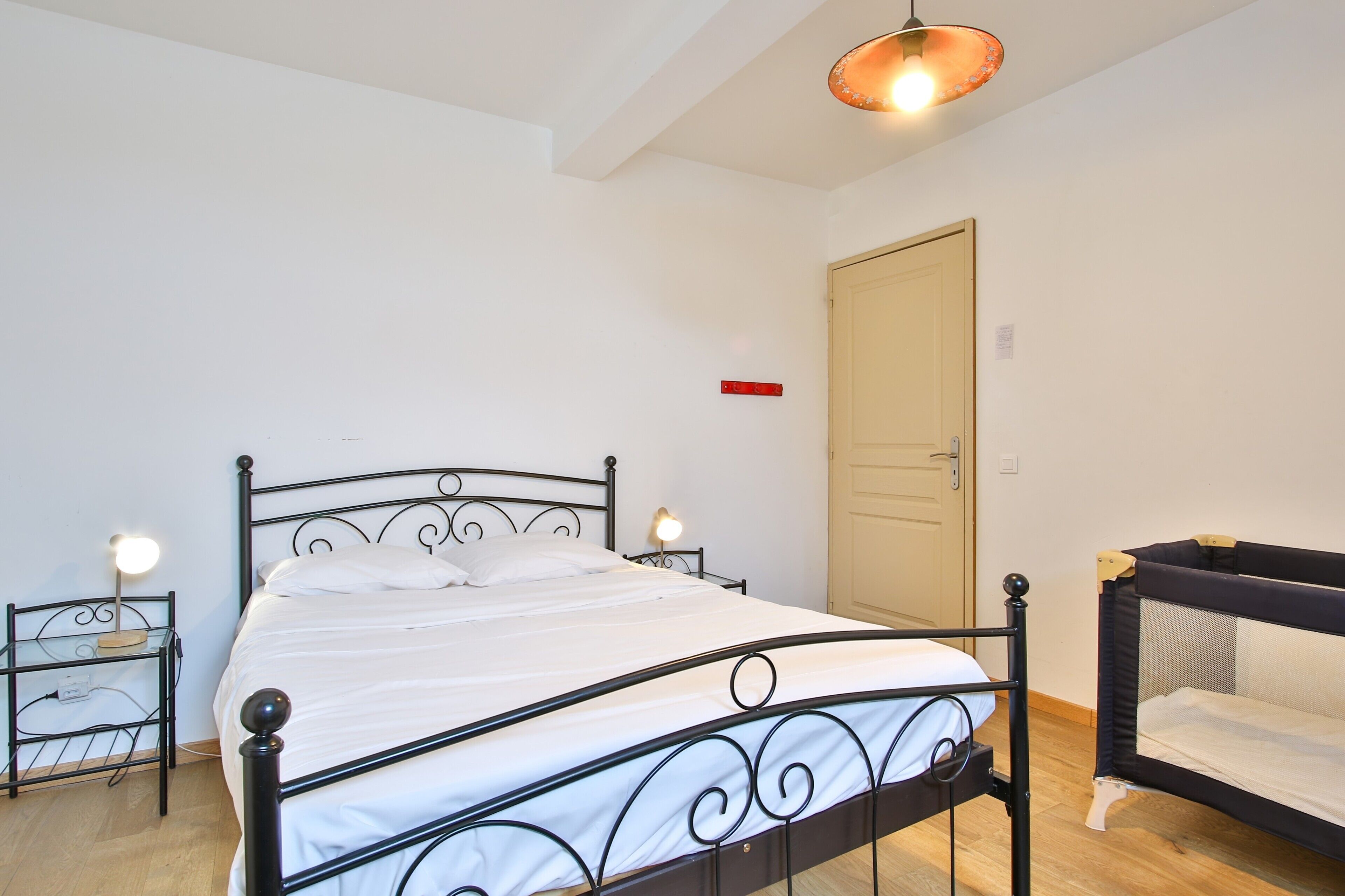 Een eenpersoonsbed met metalen frame en witte beddengoed in een kamer met witte wanden, een houten vloer en een deur met een rood bordje.