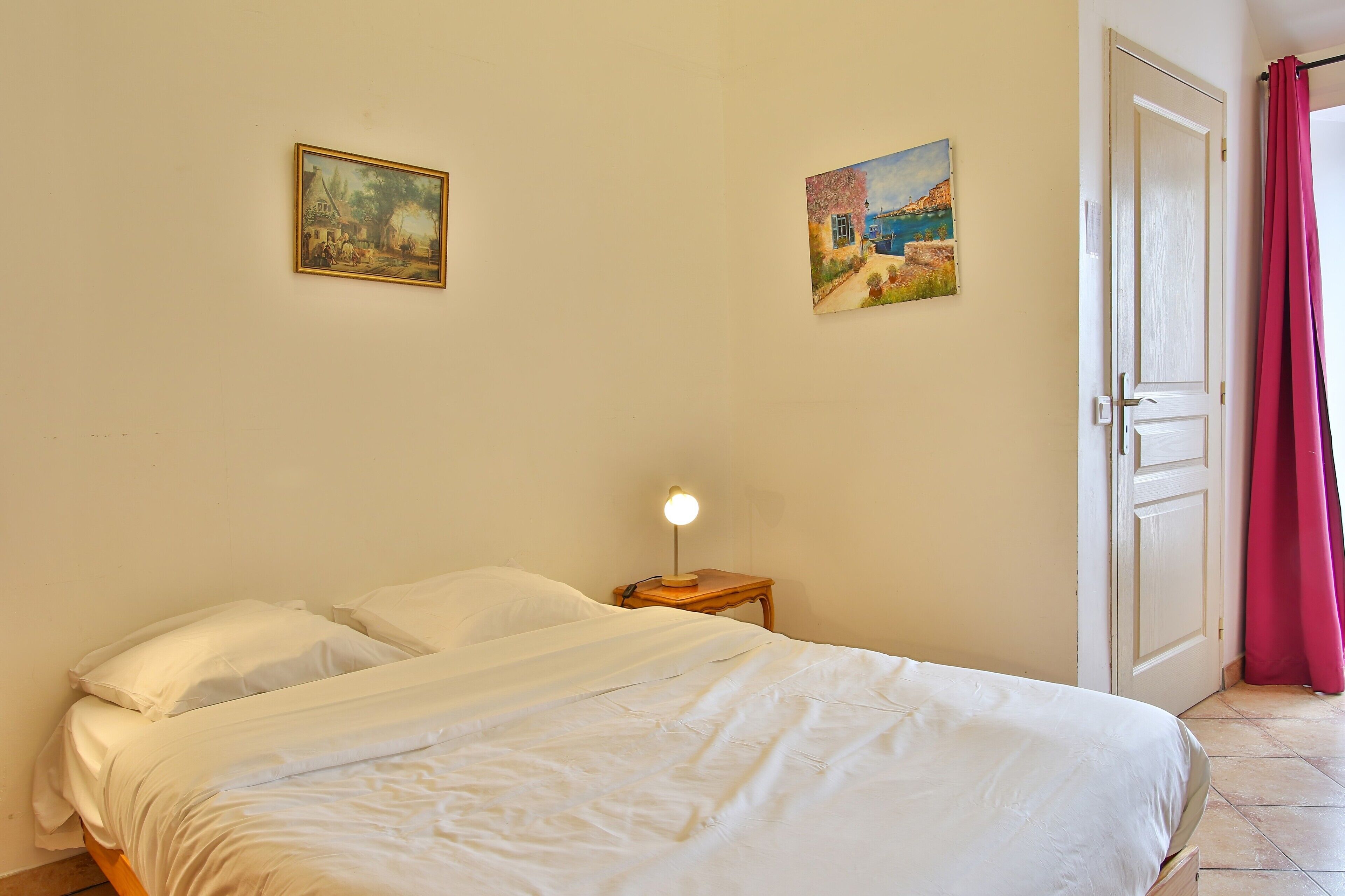 A bedroom with a bed, two paintings, a nightstand with a lamp, and a door with curtains.