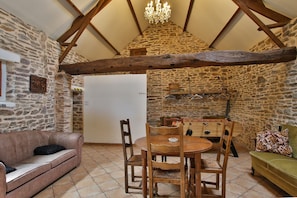 A rustic room with a stone wall, wooden beams, a chandelier, a dining table with chairs, a sofa, and a futsal table.