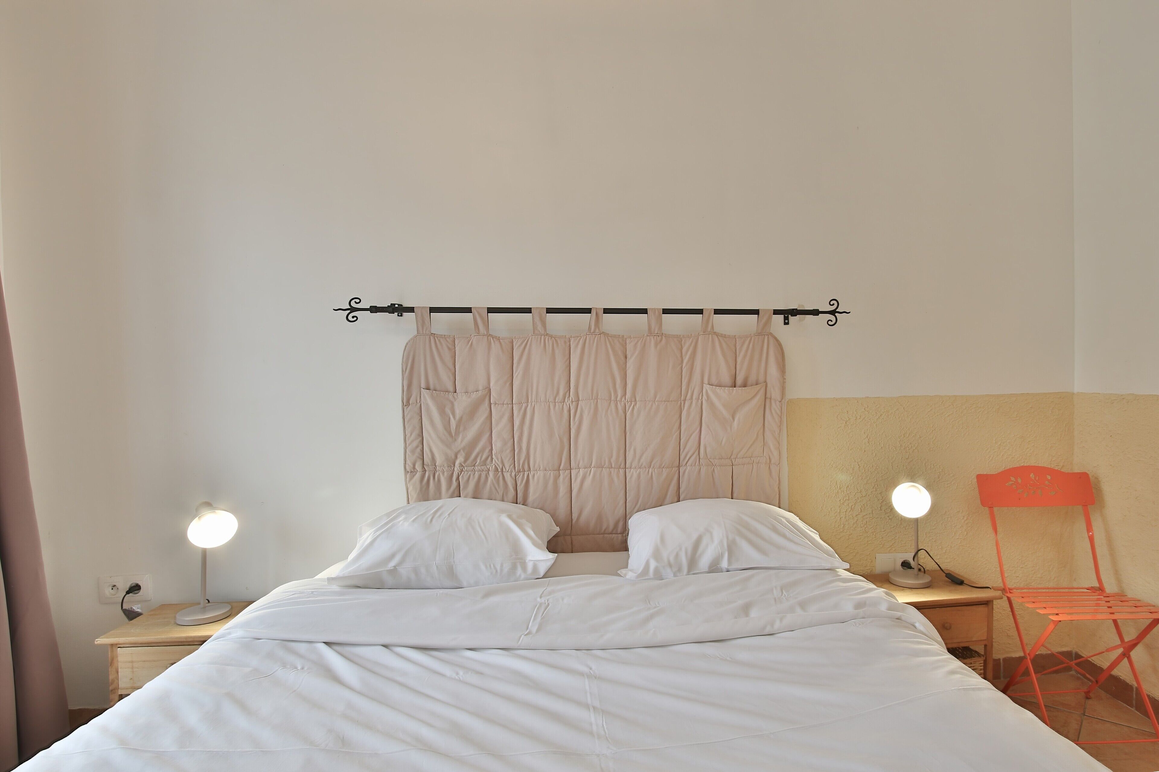 A bed with white bedding, two bedside lamps, a headboard with a curtain, and a red chair.