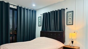 A bedroom with a bed, dark curtains, a bedside table with a lamp, and framed pictures on the wall. - Rose Park Resort (Phang Khon)