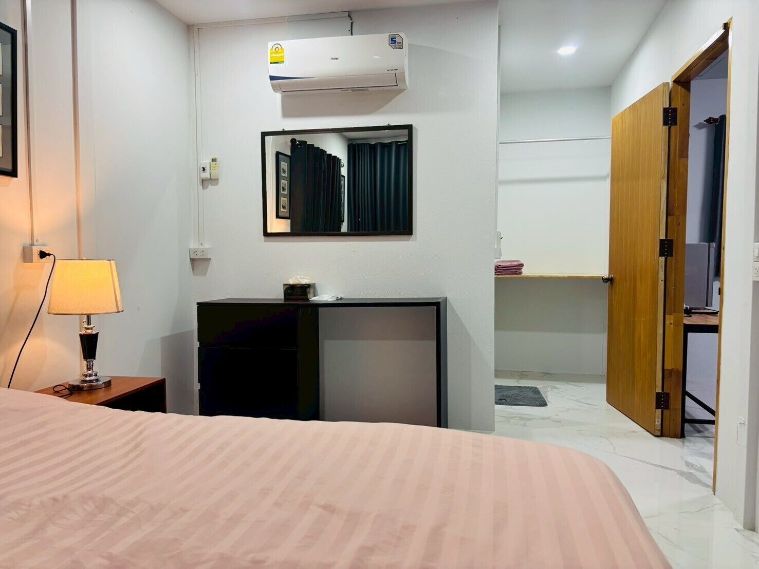 A hotel room with a bed, a desk with a lamp, an air conditioner, a mirror, and a wardrobe.