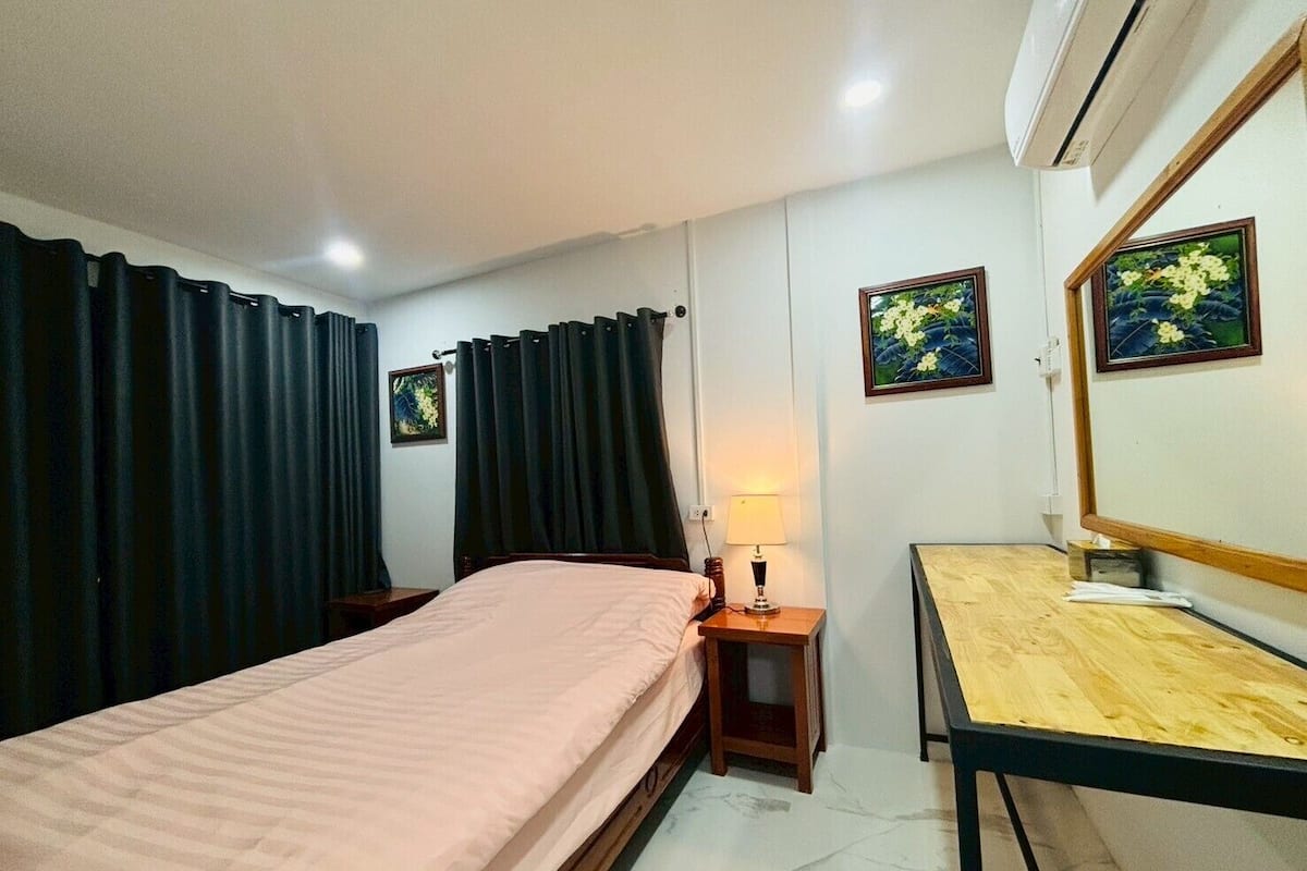 A bedroom with a bed, a bedside table with a lamp, a wooden desk, and two framed pictures on the wall.
