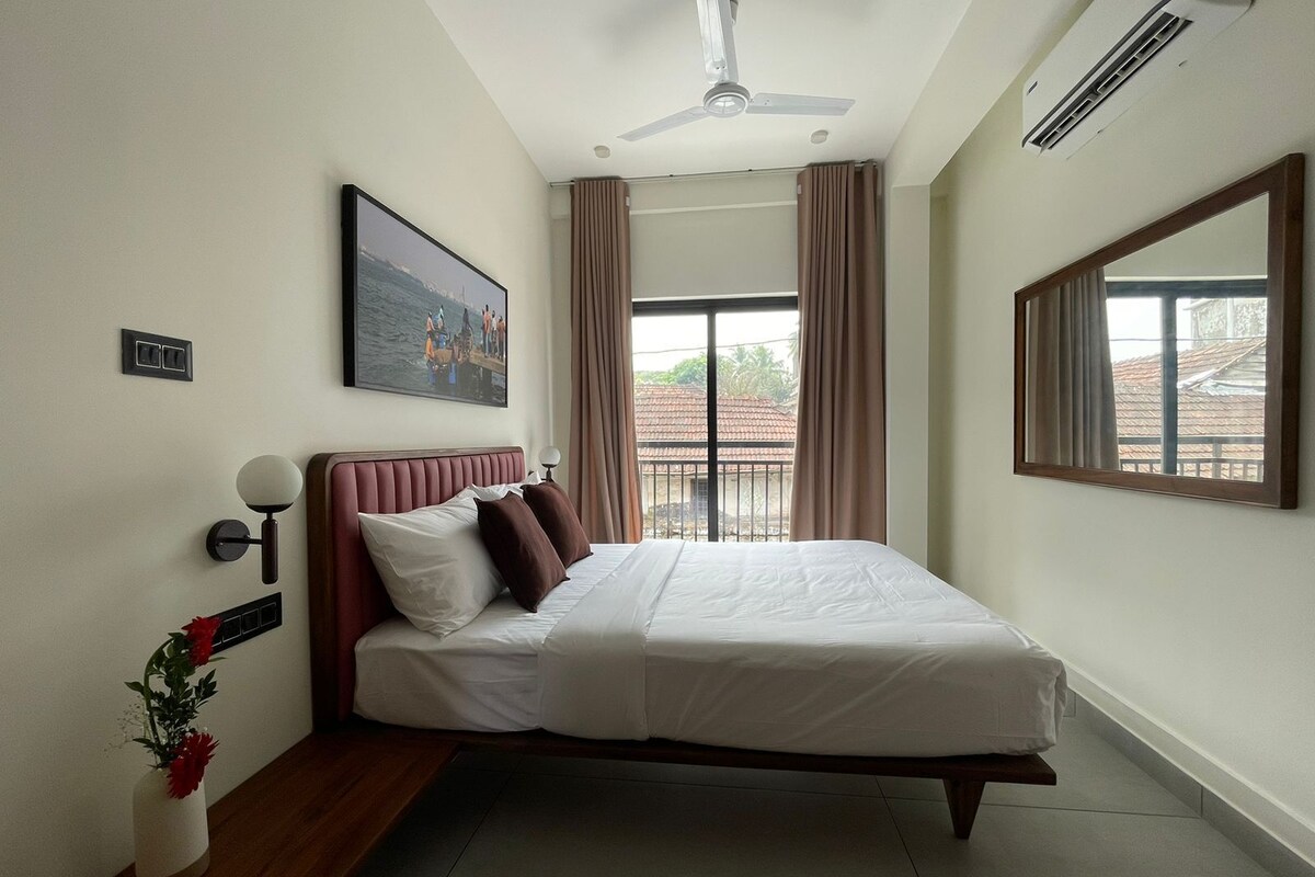 A bedroom with a bed, a ceiling fan, an air conditioning unit, a window with curtains, and a painting on the wall.