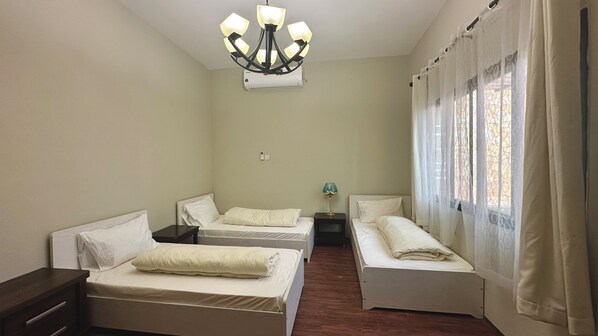 A room with two single beds, a small table with a lamp, a ceiling light fixture, and a window with curtains. - Roushan Resort (Mirbat)