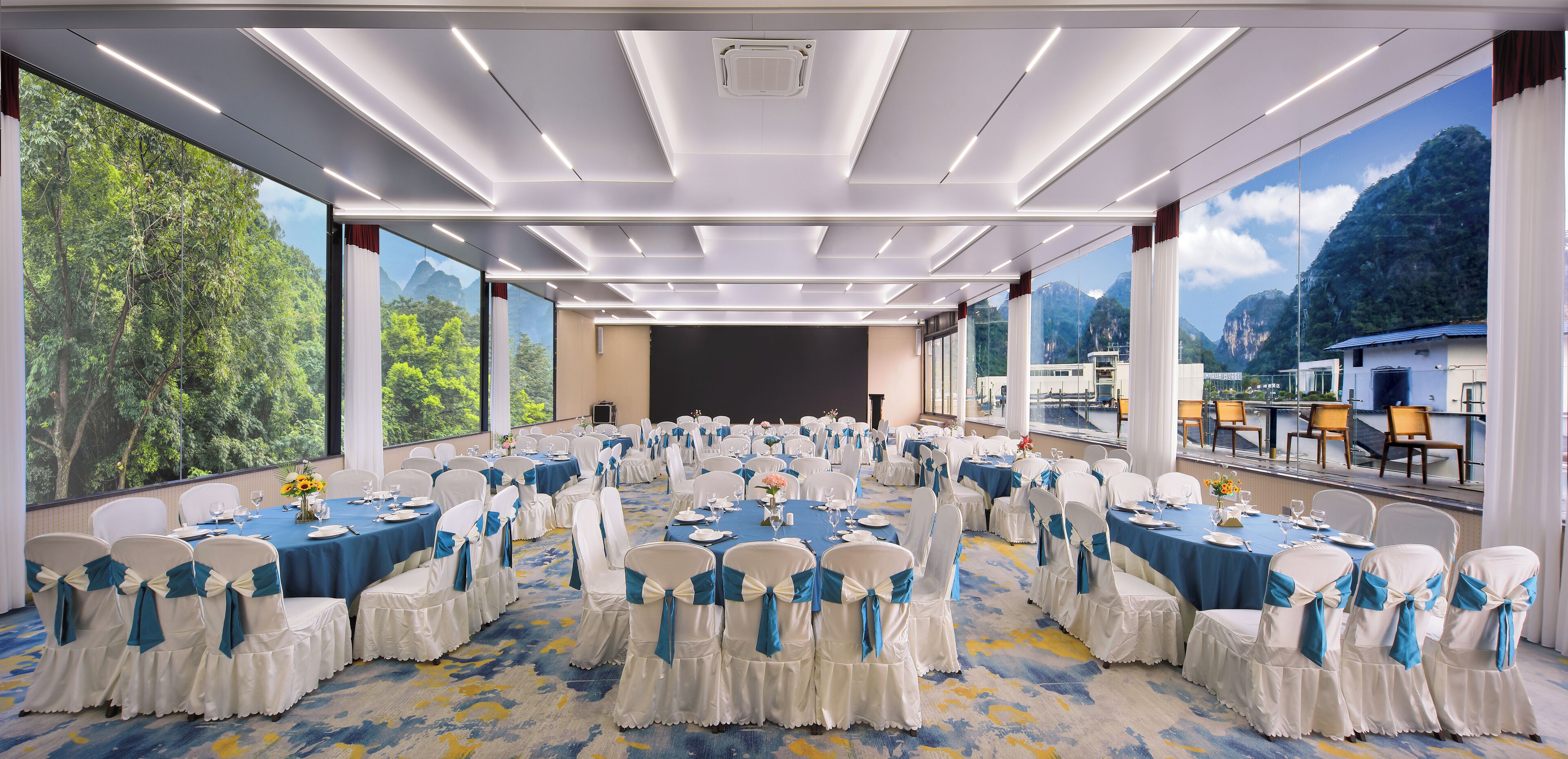 A banquet hall with round tables, white chairs with blue sashes, and large windows offering a view of a mountainous landscape.