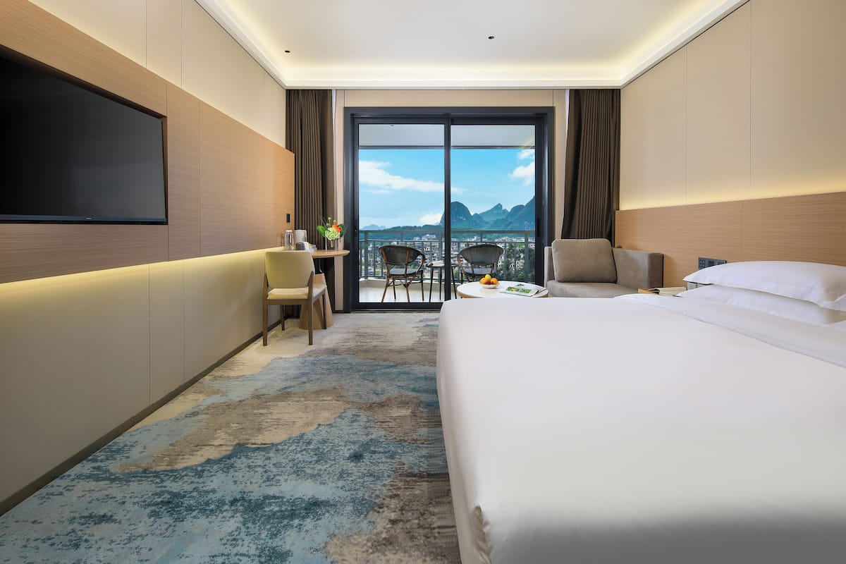 A hotel room with a large bed, a desk with a chair, a TV mounted on the wall, and a balcony with a view of mountains.