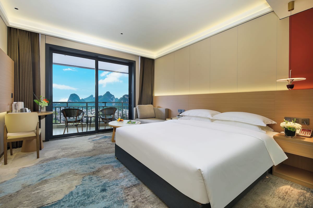 A hotel room with a large bed, a desk, a chair, and a view of the mountains through the window.