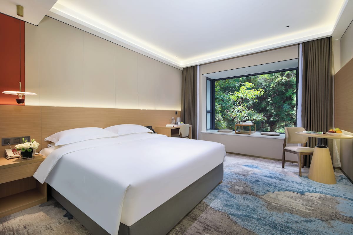 A modern hotel room with a large bed, bedside table with a lamp, a desk with a chair, and a large window offering a view of greenery.