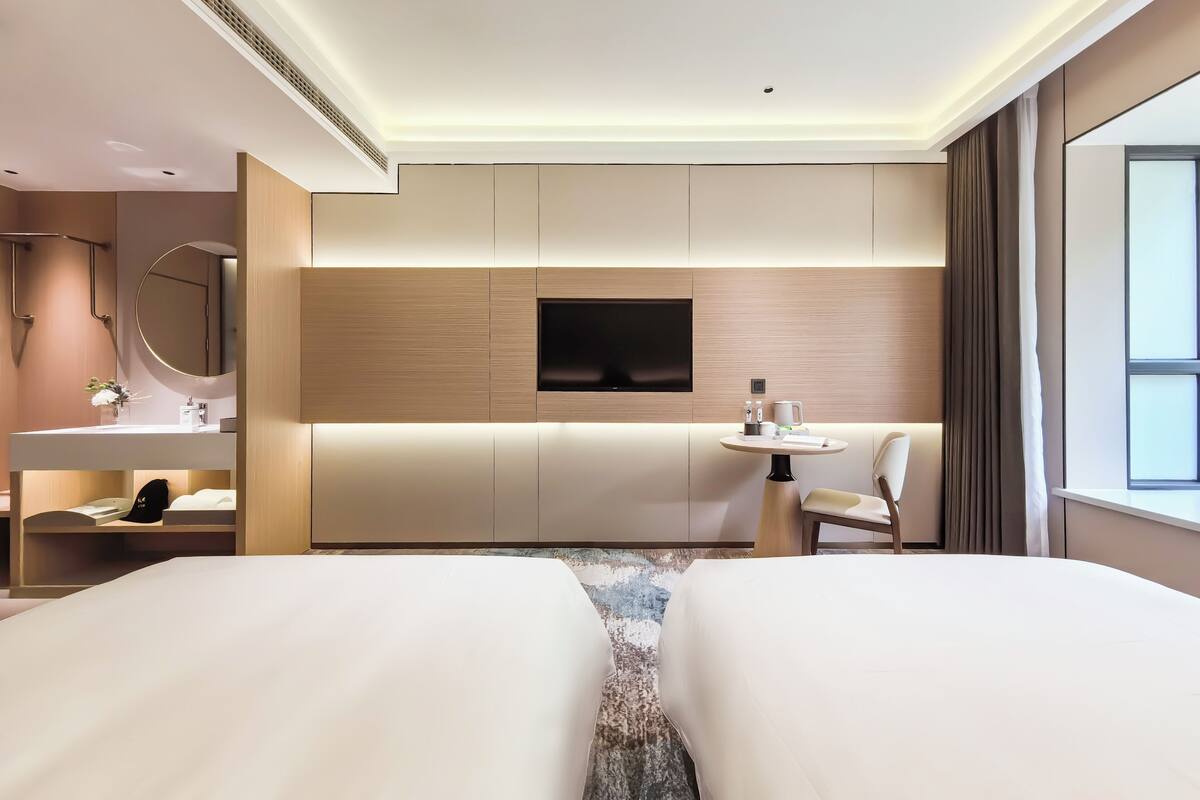 A modern hotel room with a large bed, a desk with a chair, a round mirror, a TV, and a small table with a chair.