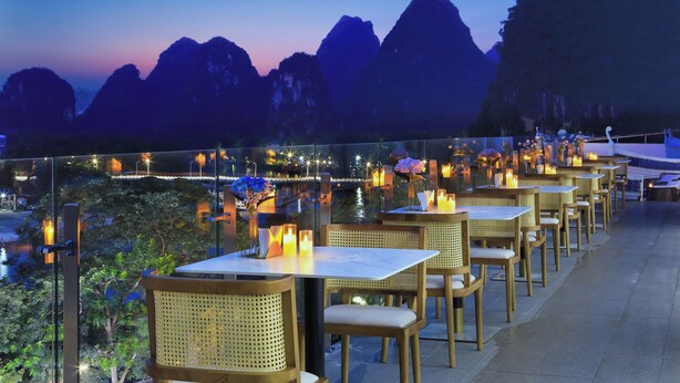 A terrace with tables and chairs, overlooking a body of water and mountains at dusk. - Langyun Garden Hotel (Guilin)