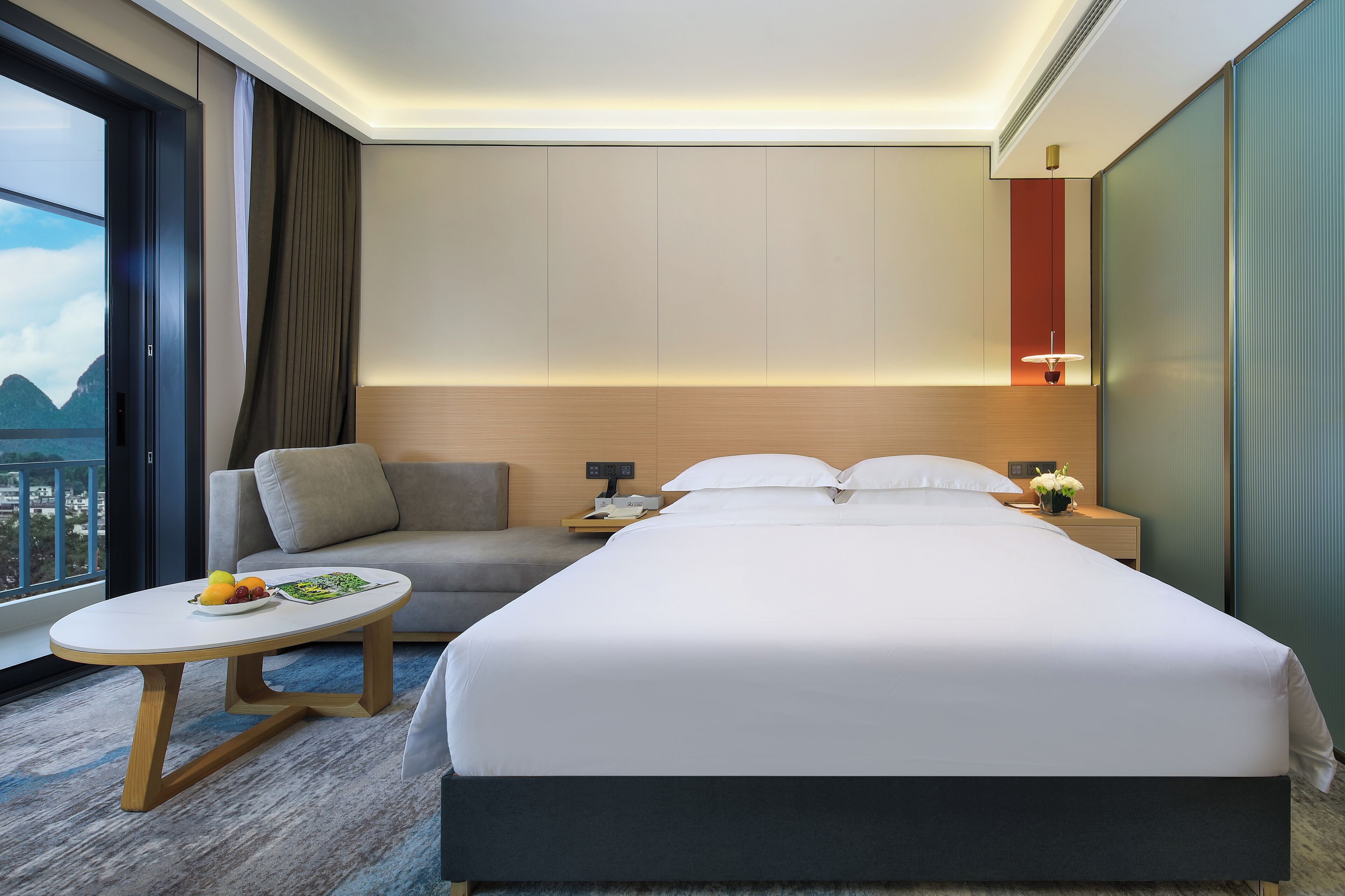 A modern hotel room with a large bed, a sofa, a small table with fruit, and a view of mountains.