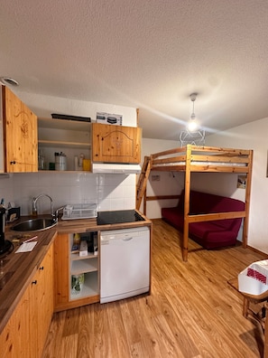 Cocina compacta con armarios de madera, lavavajillas empotrado, un lavabo y una cama nido con colchón rojo.
