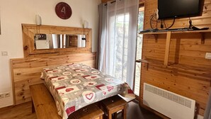 A wooden room with a bed, a table with a patterned tablecloth, a television on the wall, and a window with curtains. - Apartment "La Cabane" in Chamrousse, Private Terrace, Ski-in/Ski-out (Chamrousse)