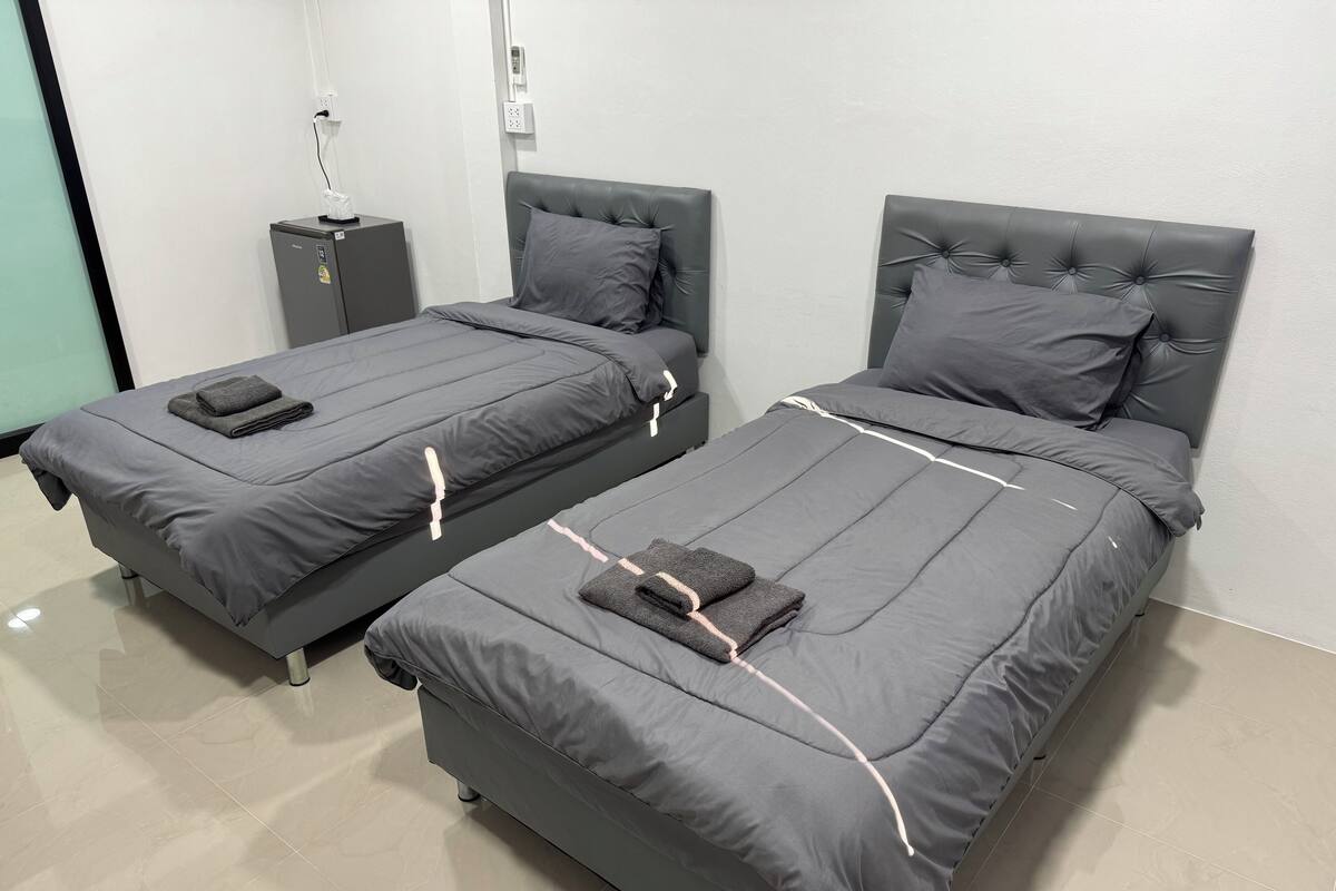 Zwei Einzelbetten mit grauer Bettwäsche und Kopfteilen, ein kleiner Nachttisch mit Lampe und eine Steckdose an der Wand.