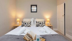 Ein ordentlich bezogenes Bett mit Kissen, ein Nachttisch mit Lampe, ein gerahmtes Bild an der Wand und eine Tür auf der rechten Seite.