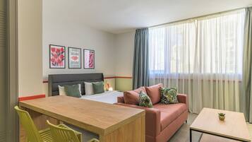 A hotel room with a bed, a sofa, a small table, and a desk. There are three framed pictures on the wall.