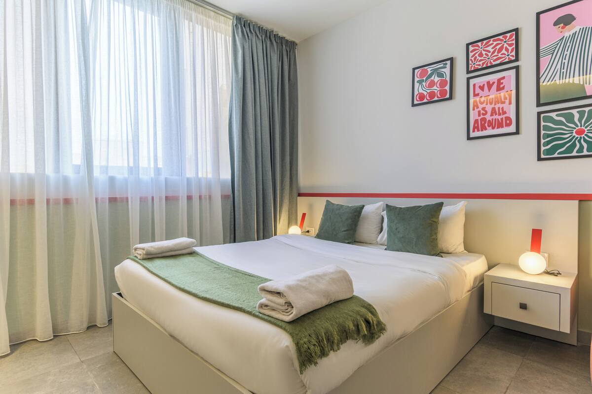 A neatly made bed with a green throw blanket, a bedside table with a lamp, and a wall with framed artwork.