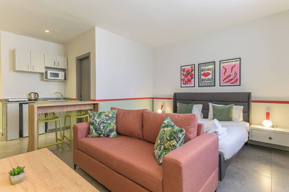 A compact hotel room with a kitchenette, a sofa with tropical cushions, a bed with a headboard, and a nightstand with a lamp.