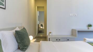 A modern hotel room with a neatly made bed, a bedside table with a lamp, a small plant, and a built-in desk with a chair.