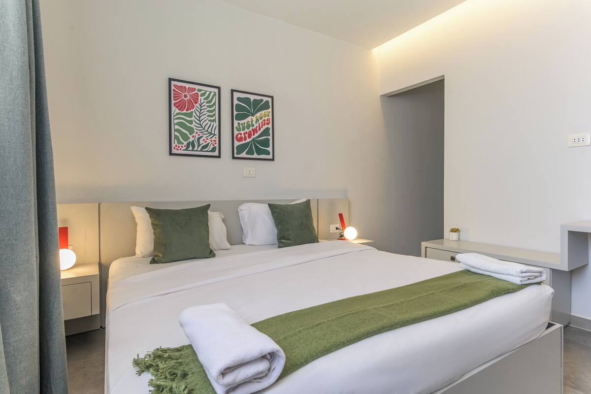 A modern hotel room with a neatly made bed, green and white bedding, two wall art pieces, a bedside table with a lamp, and a small plant on the nightstand.