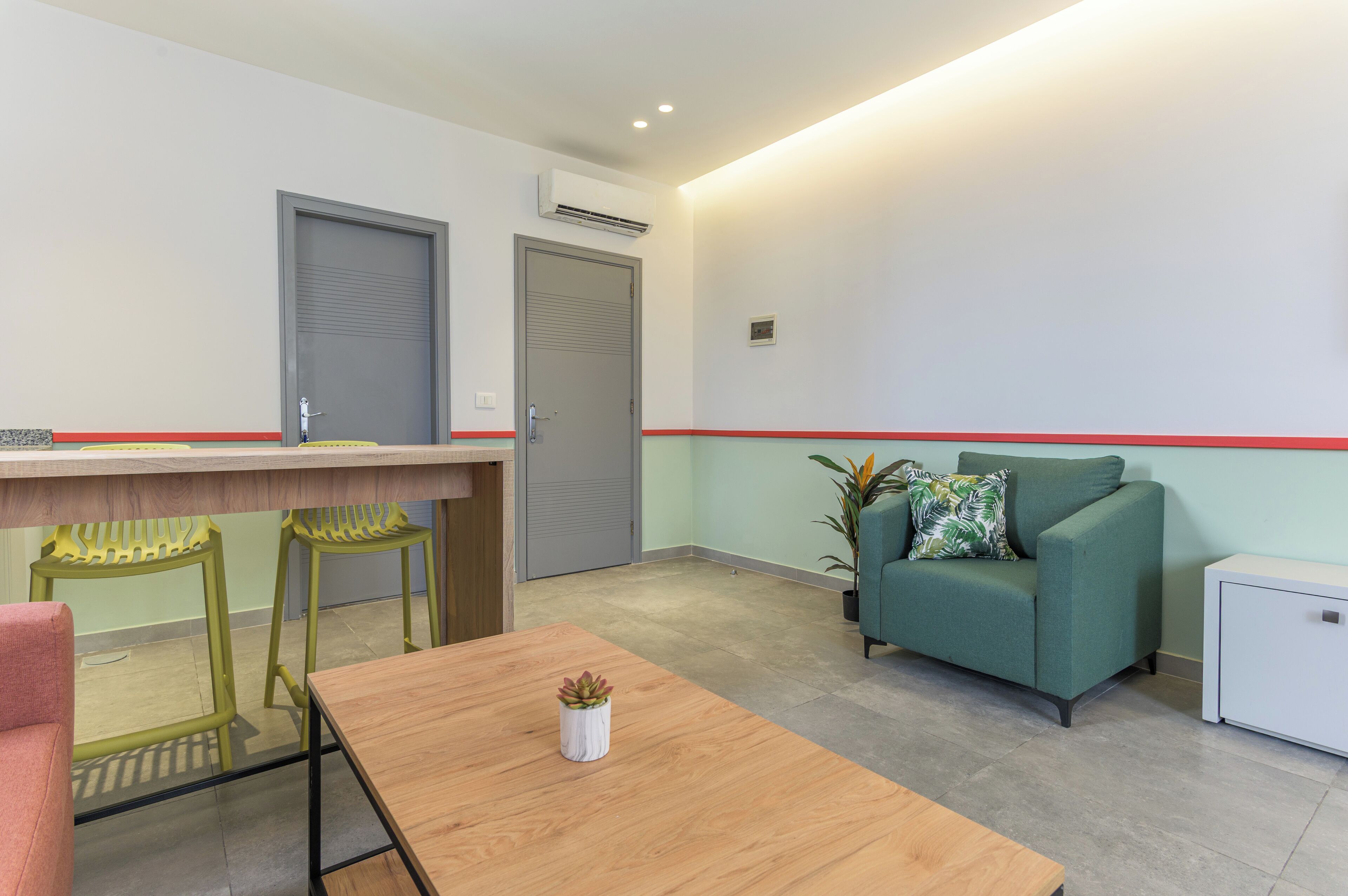 Un área de espera moderna con un mostrador de recepción, dos sillas verdes, una mesa de madera con una planta, un sofá verde y un armario blanco pequeño.