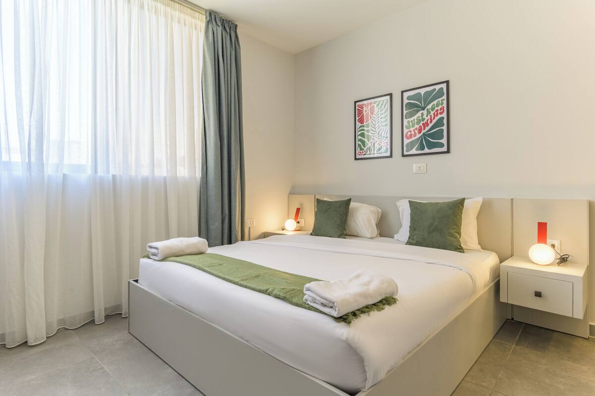 A neatly made bed with a green throw blanket, white pillows, and a white headboard. There are two bedside tables with lamps, and a wall-mounted artwork above the bed.