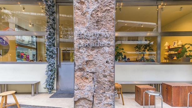 Un escaparate con un letrero que dice "the glow paradise", un pilar de piedra y asientos exteriores con taburetes de madera.