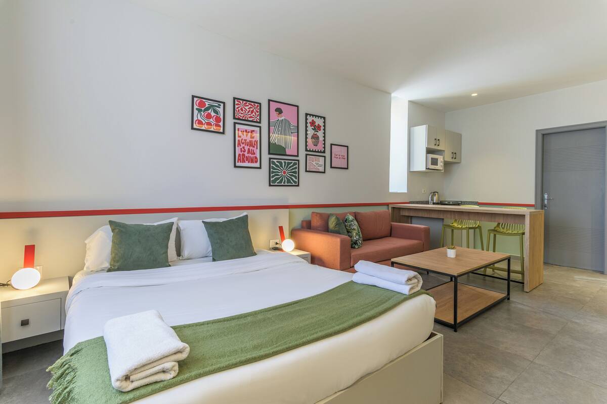 A hotel room with a bed, bedside table, lamp, and a small table with chairs. There is a kitchenette with a microwave and a door.