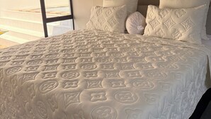 Ein großes Bett mit einer blumigen Bettdecke, ein Kopfteil mit vielen Kissen und Blick auf einen Pool durch ein Fenster.