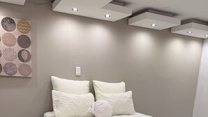 Ein ordentlich bezogenes Bett mit weißer Decke und Kissen, eine Wand mit einem runden Wandkunstwerk und Einbaustrahler.