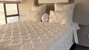 Ein großes Bett mit weißer Steppdecke, ein Fenster mit Jalousien und eine schlichte Wand.