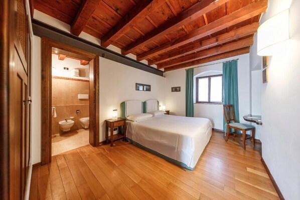 A bedroom with a wooden ceiling, a bed, a nightstand, a chair, and a window with curtains. - Serego Alighieri (Sant'Ambrogio di Valpolicella)