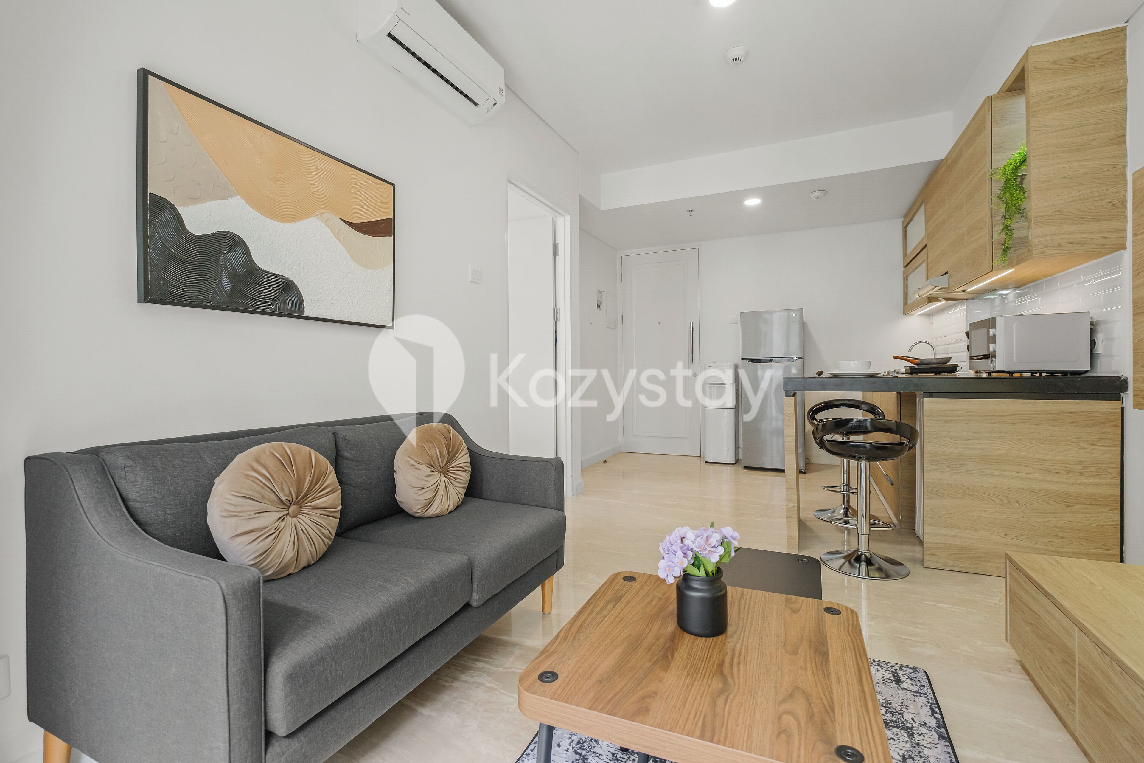 A modern living room with a grey sofa, a wooden coffee table with a vase of flowers, and a kitchen area in the background.